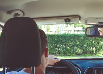 View from the backseat of a car, a young man is driving, can see his smiling face in the rearview mirror.