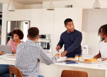 Family of five sitting or standing around island countertop, talking and preparing food.