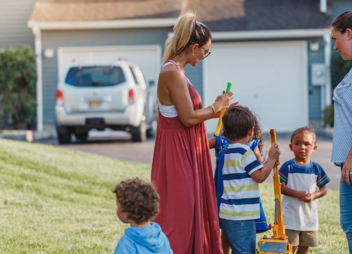 Women and children talk outside in front of homes and a grassy yard